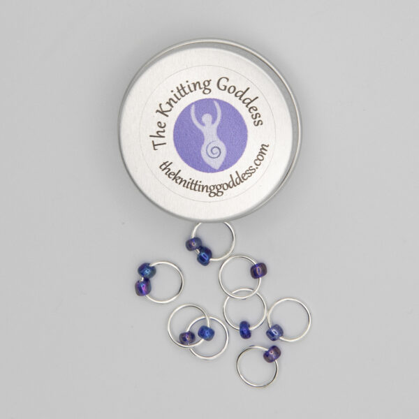 set of eight stitch markers made with jump rings and blue beads. Shown with a small round storage tin with The Knitting Goddess logo