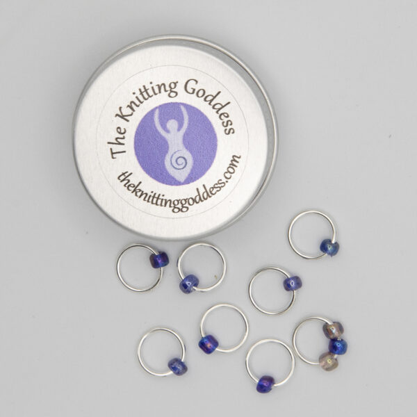 set of eight stitch markers made with jump rings and blue and purple beads. Shown with a small round storage tin with The Knitting Goddess logo
