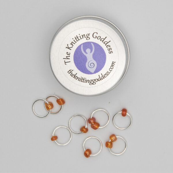 set of eight stitch markers made with jump rings and orange beads. Shown with a small round storage tin with The Knitting Goddess logo