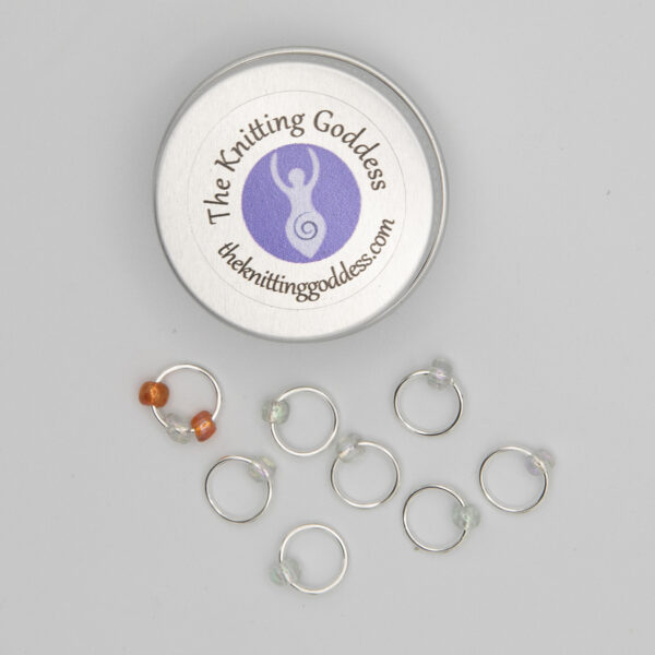 set of eight stitch markers made with jump rings and pearl and orange beads. Shown with a small round storage tin with The Knitting Goddess logo