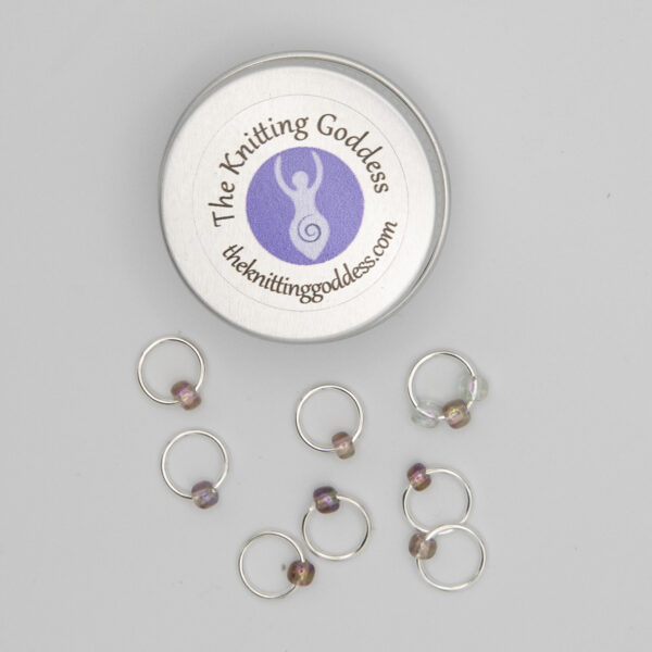 set of eight stitch markers made with jump rings and purple beads. Shown with a small round storage tin with The Knitting Goddess logo