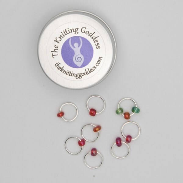 set of eight stitch markers made with jump rings and red and green beads. Shown with a small round storage tin with The Knitting Goddess logo