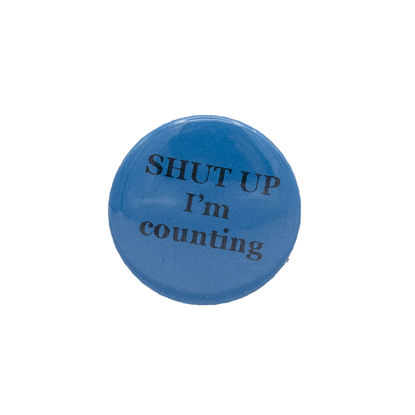 Turquoise button badge with black writing which reads SHUT UP I'm counting