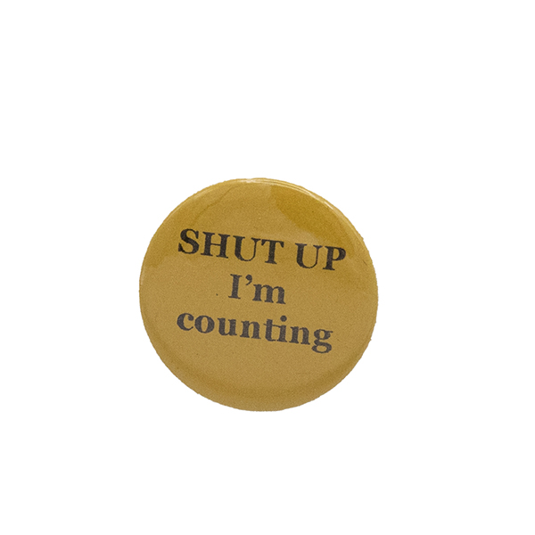 Yellow button badge with black writing which reads SHUT UP I'm counting