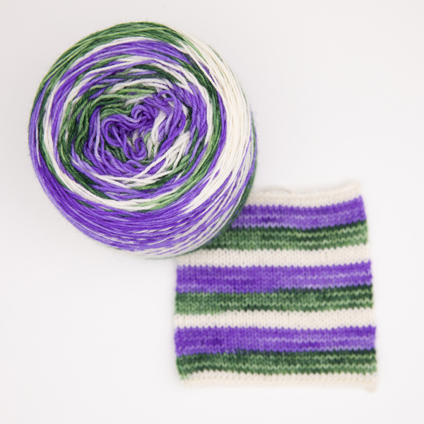 Overhead view. Ball of gender queer pride flag self striping sock yarn. Yarn is dyed in the the three colours of the flag - olive green, white and purple. Shown with a knitted sample showing how the yarn will stripe into 4-5 rounds of each colour.