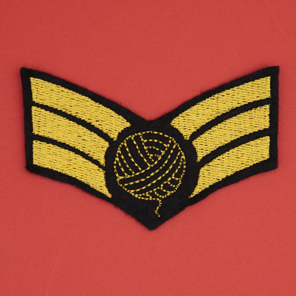 embroidered patch on black felt, embroidery in golden yellow. Design is three chevron stripes with a ball of yarn on top.. shown on a red background