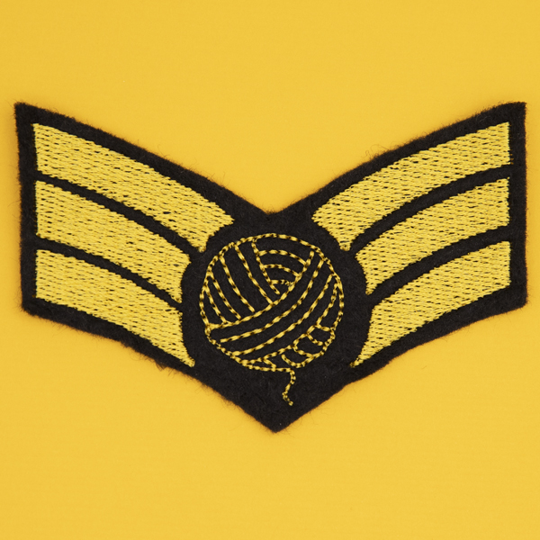 embroidered patch on black felt, embroidery in golden yellow. Design is three chevron stripes with a ball of yarn on top..yellow background