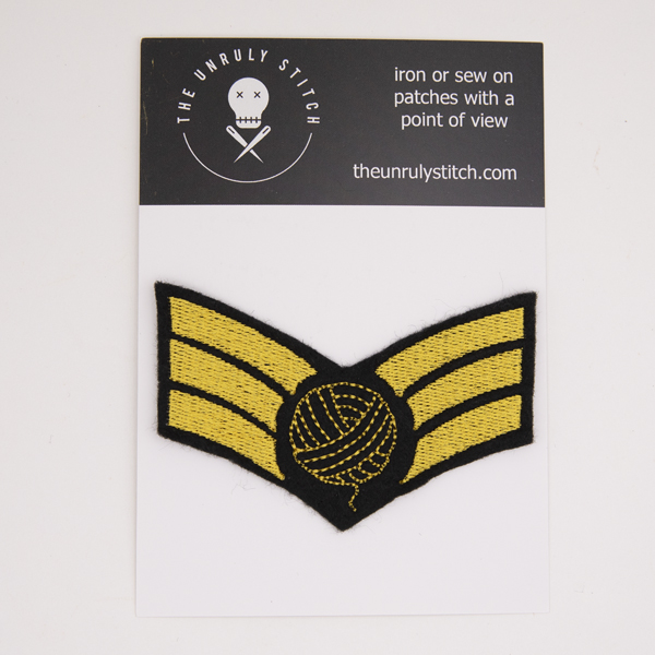 embroidered patch on black felt, embroidery in golden yellow. Design is three chevron stripes with a ball of yarn on top... shown on a postcard which has instructions on the back