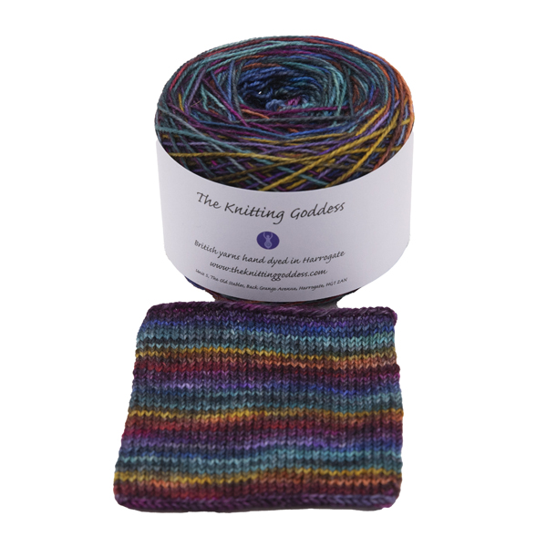 A yarn cake with The Knitting Goddess ball band. Yarn is hand dyed in rainbow colours and overdyed with black. SWatch shows how the yarn knits up in stripes f one or two rounds of a sock.