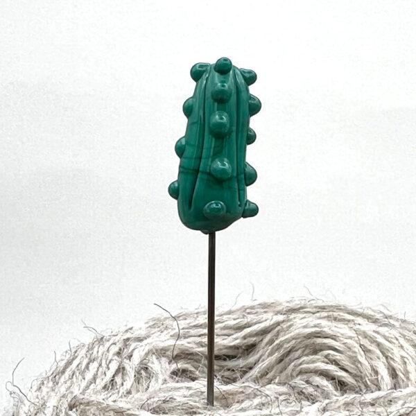 green glass pickle pin with point stuck into a ball of yarn so the shaft of the pin is visible