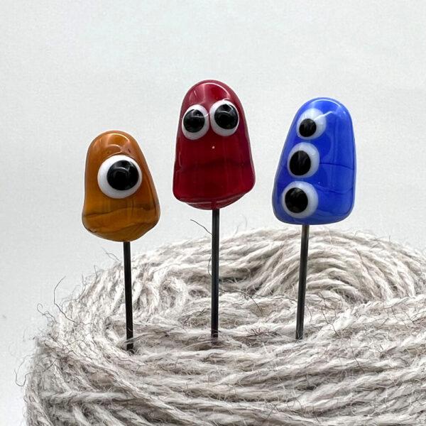 three little monster pins with googly eyes, points stuck into ball of yarn so the shaft of the pins is visible