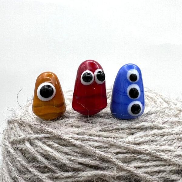 three little monster pins with googly eyes, stuck into ball of yarn. Monsters are gold with one eye, red with two eyes and blue with three eyes