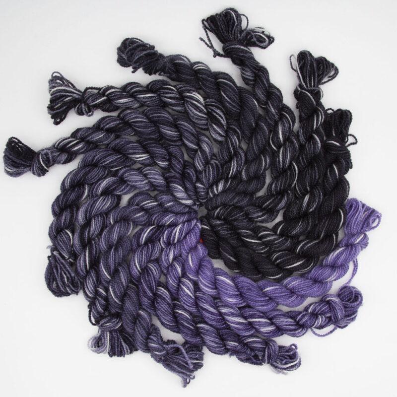 set of 12 mini skeins in all the stars in the sky colourway. Skeins go from purple to black and all have white splodges. Arranged in a spiral. Shot from above.