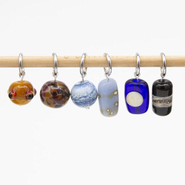 set of six stitch markers - three round (yellow with red and black dots, purple and orange speckles on grey, blue and white swirl), 3 column shaped (pale blue with silver dots, dark blue with cream moon. black with cream and silver swirl). The markers have jump ring findings.