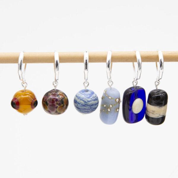 set of six stitch markers - three round (yellow with red and black dots, purple and orange speckles on grey, blue and white swirl), 3 column shaped (pale blue with silver dots, dark blue with cream moon. black with cream and silver swirl). The markers have latch back hooks