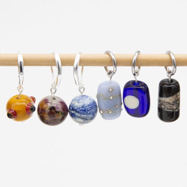 set of six stitch markers - three round (yellow with red and black dots, purple and orange speckles on grey, blue and white swirl), 3 column shaped (pale blue with silver dots, dark blue with cream moon. black with cream and silver swirl). The round stitch markers have latch back hooks, the rest have jump ring findings.