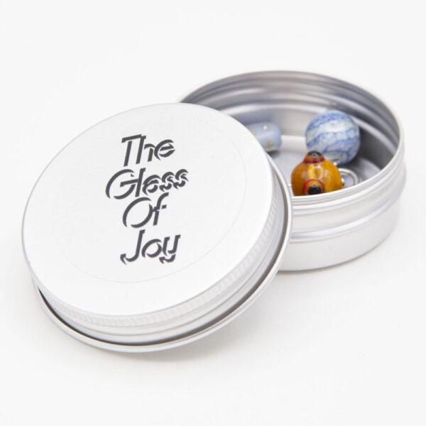 round metal tin with The Glass Of Joy logo. The tin is open with the crew top lid resting on the side, and several stitch markers are visible inside the tin