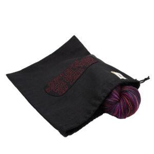 black linen project bag embroidered with "YOU KNOW IT'S LOVE WHNE YOU WOULD KNIT THEM BLACK SOCKS" in red outline text. Bag closes with a drawstring and has The Unruly Stitch label. Bag is open and the top of a skein of True Love yarn (red, black and pink) shows.