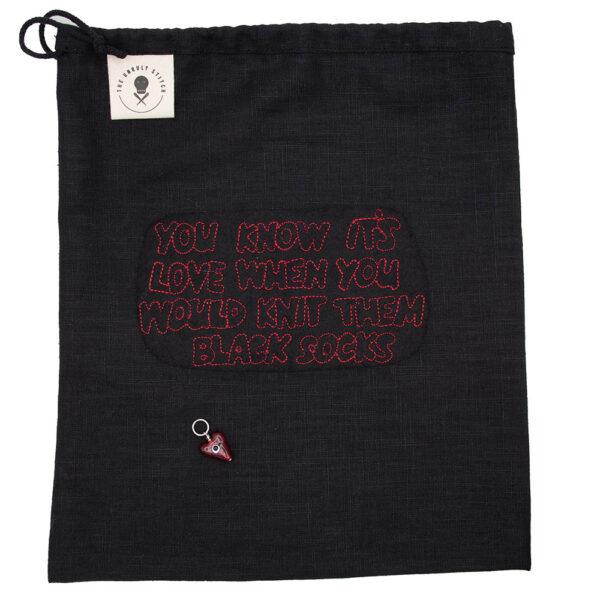 flat view of black linen project bag embroidered with "YOU KNOW IT'S LOVE WHNE YOU WOULD KNIT THEM BLACK SOCKS" in red outline text. Bag closes with a drawstring and has The Unruly Stitch label. Glass heart stitch marker sits on bag.