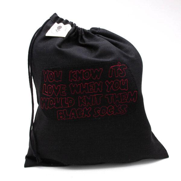 black linen project bag embroidered with "YOU KNOW IT'S LOVE WHNE YOU WOULD KNIT THEM BLACK SOCKS" in red outline text. Bag closes with a drawstring and has The Unruly Stitch label.