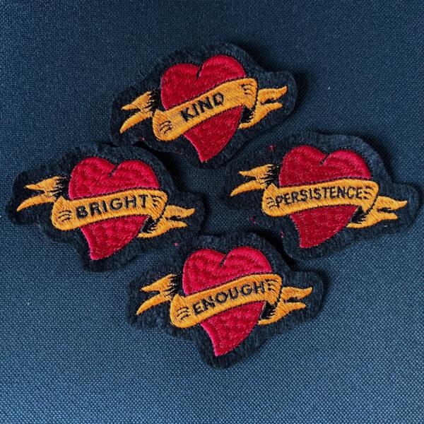four red tattoo style embroidered hearts with orange banner and text "KIND", "BRIGHT", "PERSISTENCW" and "ENOUGH"