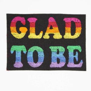 Embroidered patch with text "GLAD TO BE" the background is black felt and the letters are embroidered with stripes of colour to form an eight colour pride flag.