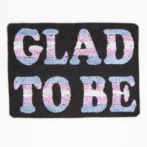 Embroidered patch with text "GLAD TO BE" the background is black felt and the letters are embroidered with stripes of colour to the trans pride flag.