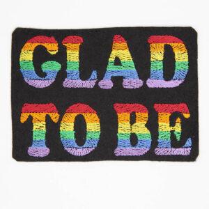Embroidered patch with text "GLAD TO BE" the background is black felt and the letters are embroidered with stripes of colour to form a six colour rainbow.